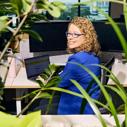 A woman with curly hair and glasses, wearing a blue blazer, sits at a desk with a laptop. She's smiling, surrounded by green plants in an office.