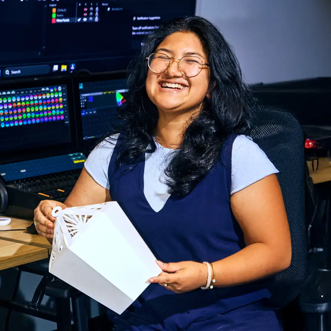A woman with long dark hair and glasses smiles joyfully while holding a white geometric object. She sits by a desk with colorful computer screens.