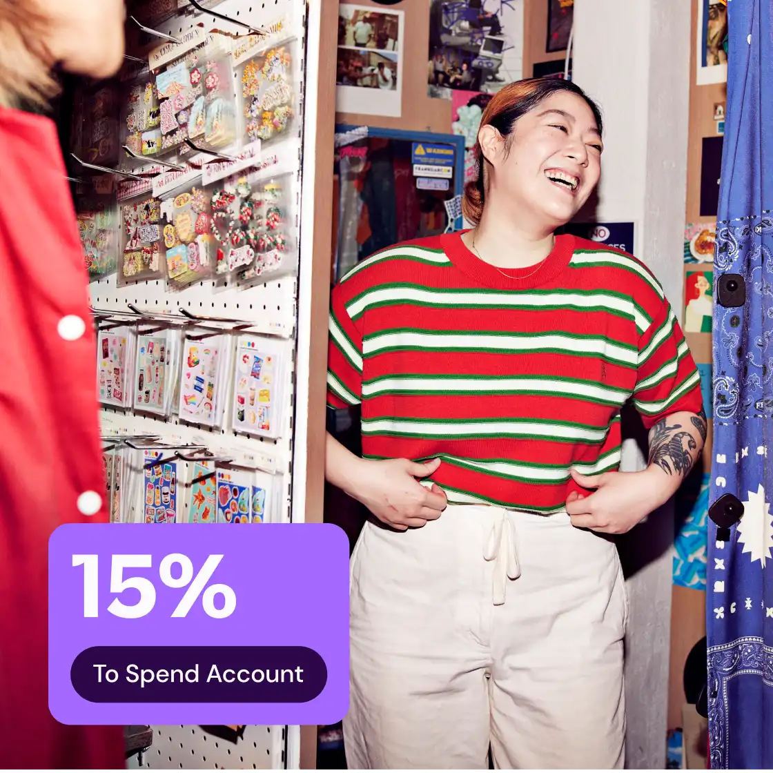 Person laughing in a colorful store, wearing a red and green striped shirt. A "15% To Spend Account" promo is displayed on the image.