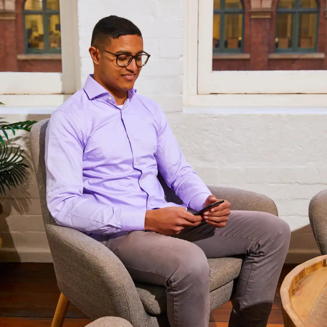 A person in a lavender shirt sits in a gray armchair holding a smartphone, smiling. They are indoors, near a window, creating a relaxed atmosphere.