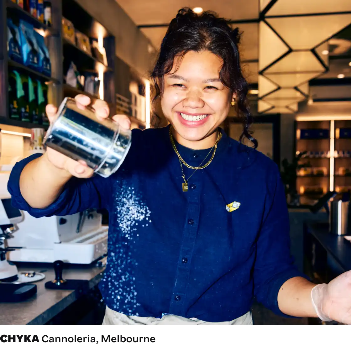 Smiling woman in a blue shirt joyfully sprinkles sugar from a metal shaker in a bustling, well-lit cafe. The atmosphere is warm and inviting.