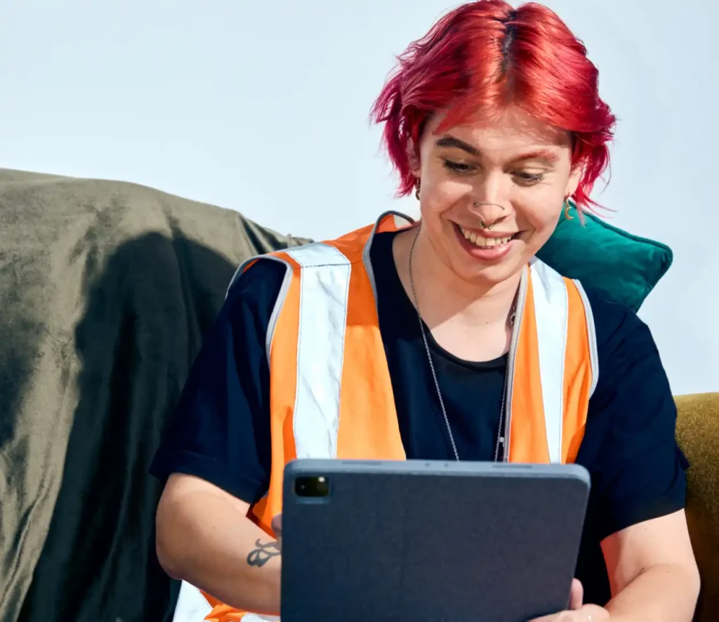 Person with short red hair and a nose ring smiles while looking at a tablet. They wear an orange safety vest and sit on a couch, conveying a cheerful mood.