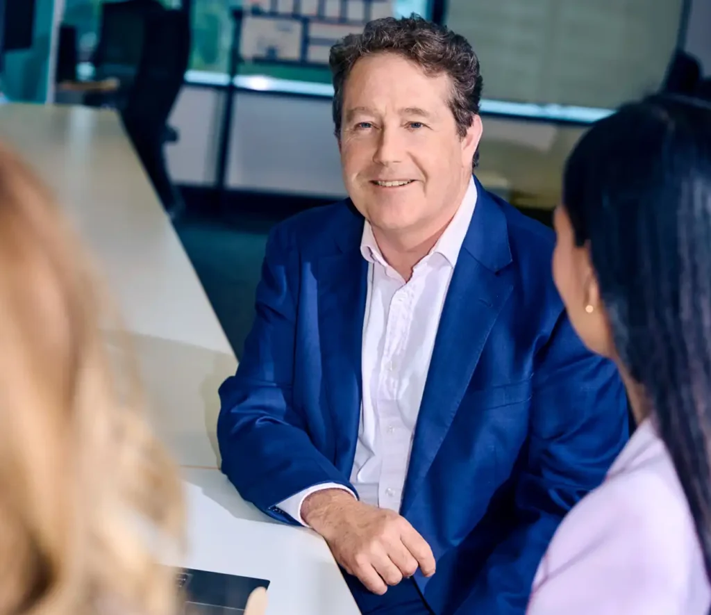 A man in a blue suit and white shirt is smiling, seated at a conference table with two women. The setting conveys a professional and friendly atmosphere.