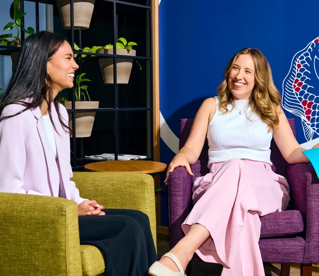 Two women sit in colorful chairs, smiling and conversing. The setting is vibrant, with potted plants on shelves and blue walls in the background.