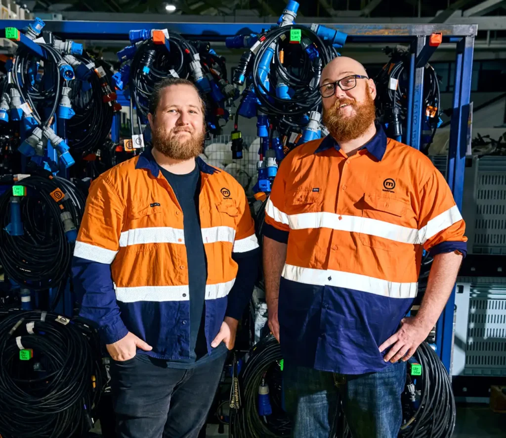 Two men in orange safety shirts stand in front of electrical cables. They appear confident and content, suggesting a work environment focused on safety and teamwork.