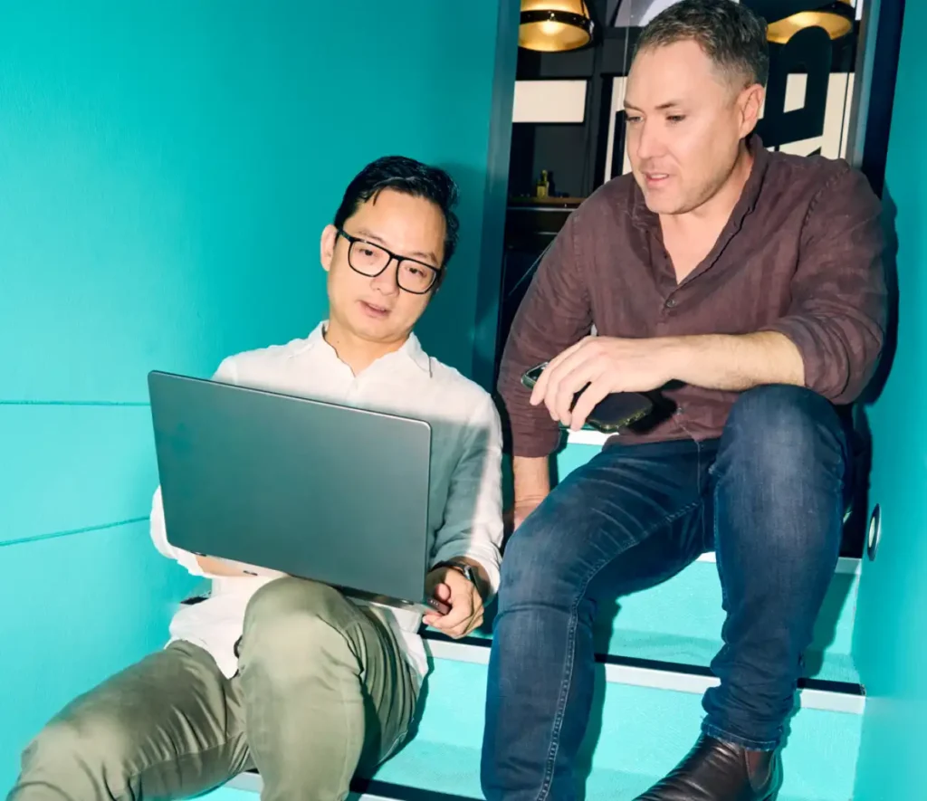 Two men sit on turquoise stairs, engaged with a laptop. The atmosphere is casual and collaborative, with soft lighting enhancing focus and connection.