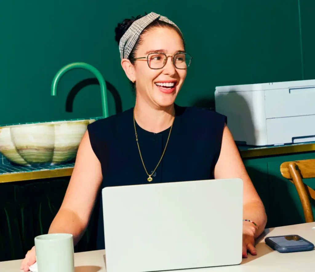 A woman with glasses and a headband smiles while using a laptop at a table. Behind her is a green wall, a ceramic sink, and a printer. Bright, cheerful atmosphere.