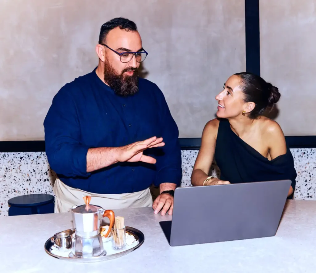 A man and woman engage in a friendly discussion at a table with a laptop and coffee set. The woman smiles, creating a collaborative ambiance.