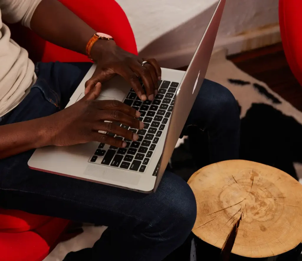 A person types on a laptop while seated on a red chair. They wear jeans and a beige sweater, with a wooden stump table nearby, creating a cozy, focused atmosphere.
