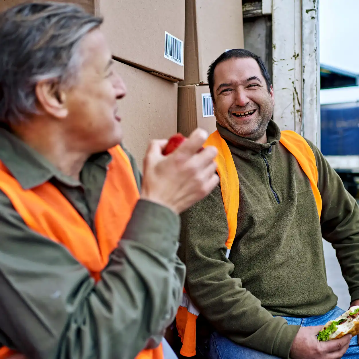 Two men wearing orange safety vests share a laugh during a break, sitting by stacked cardboard boxes. They hold sandwiches, conveying a relaxed, cheerful mood.