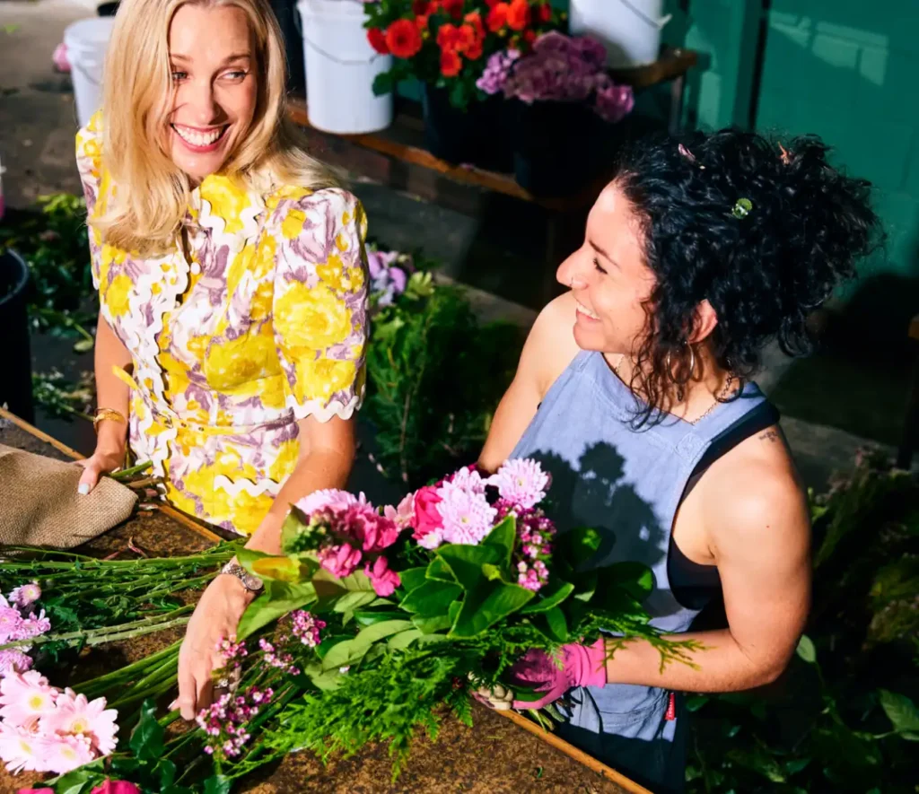 Two women in a vibrant flower shop smile at each other while arranging bouquets. One holds pink flowers, surrounded by colorful blooms and greenery.