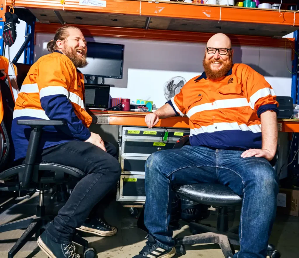 Two men in high-visibility jackets and jeans are laughing together in a workshop. They are seated near a desk with shelves, conveying a joyful mood.