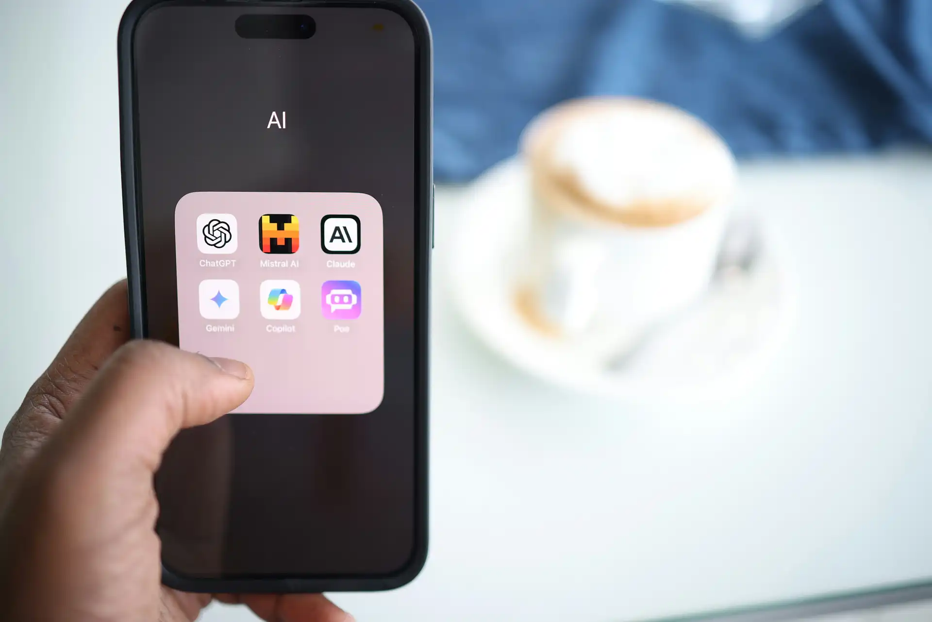 A hand holds a smartphone displaying AI app icons, including ChatGPT, Midjourney, Claude, Gemini, Copilot, and Poe. A blurred cappuccino is in the background.