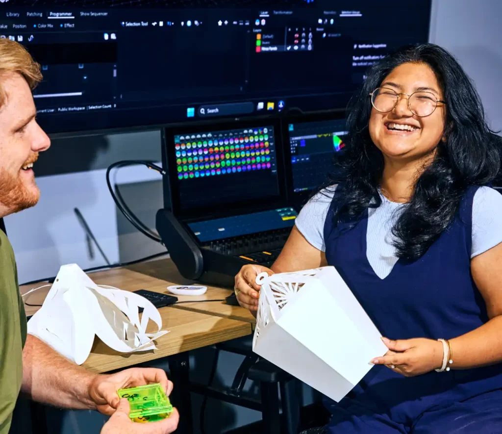 Two people laughing and crafting at a table with a computer screen displaying colorful graphics. The mood is creative and joyful.