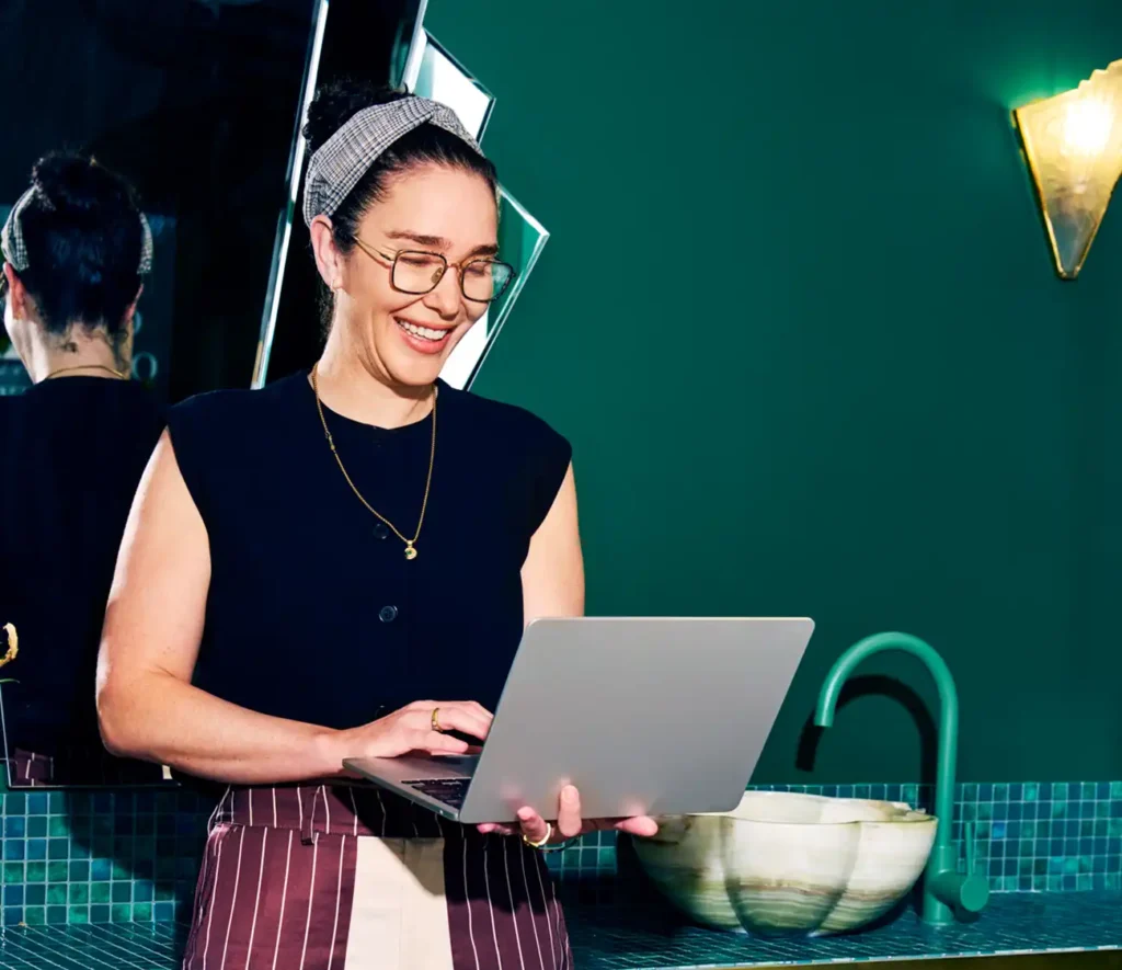 A woman with glasses, wearing a headband and apron, smiles at a laptop in a stylish kitchen with green walls and a modern sink.