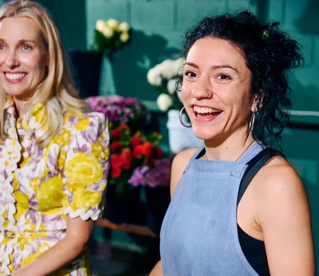 Two women are smiling in a floral shop. The woman on the left wears a yellow and white floral dress, the woman on the right wears a blue apron. Vibrant flowers in the background add a joyful tone.