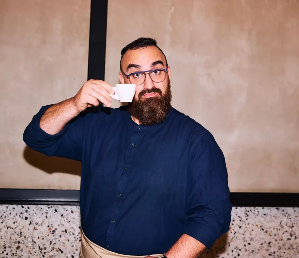 Bearded man in glasses and a navy shirt playfully holds a small white cup to his eye. Background features a textured beige wall. Playful tone.