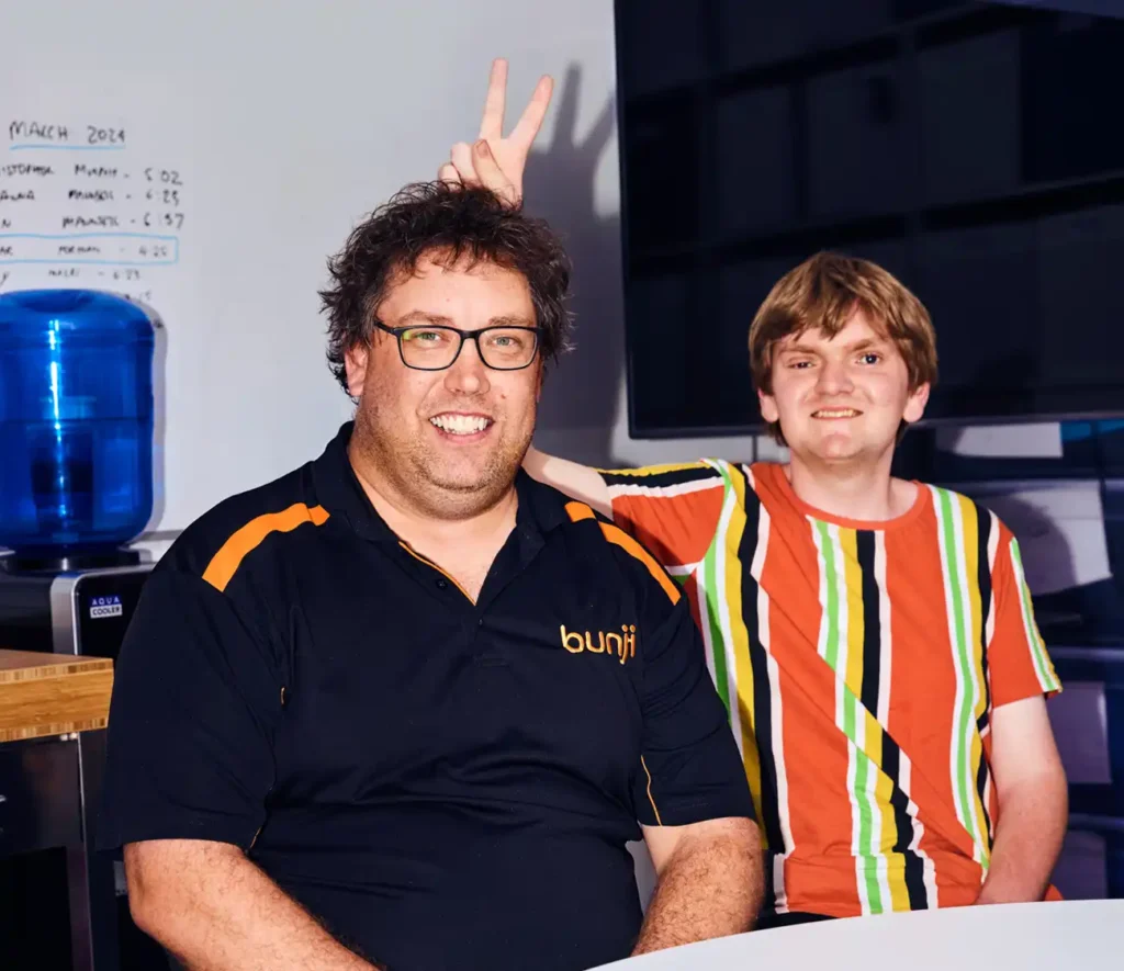 Two people sit side by side smiling in an office. The person on the right playfully gives bunny ears to the other. A water cooler and whiteboard are visible.