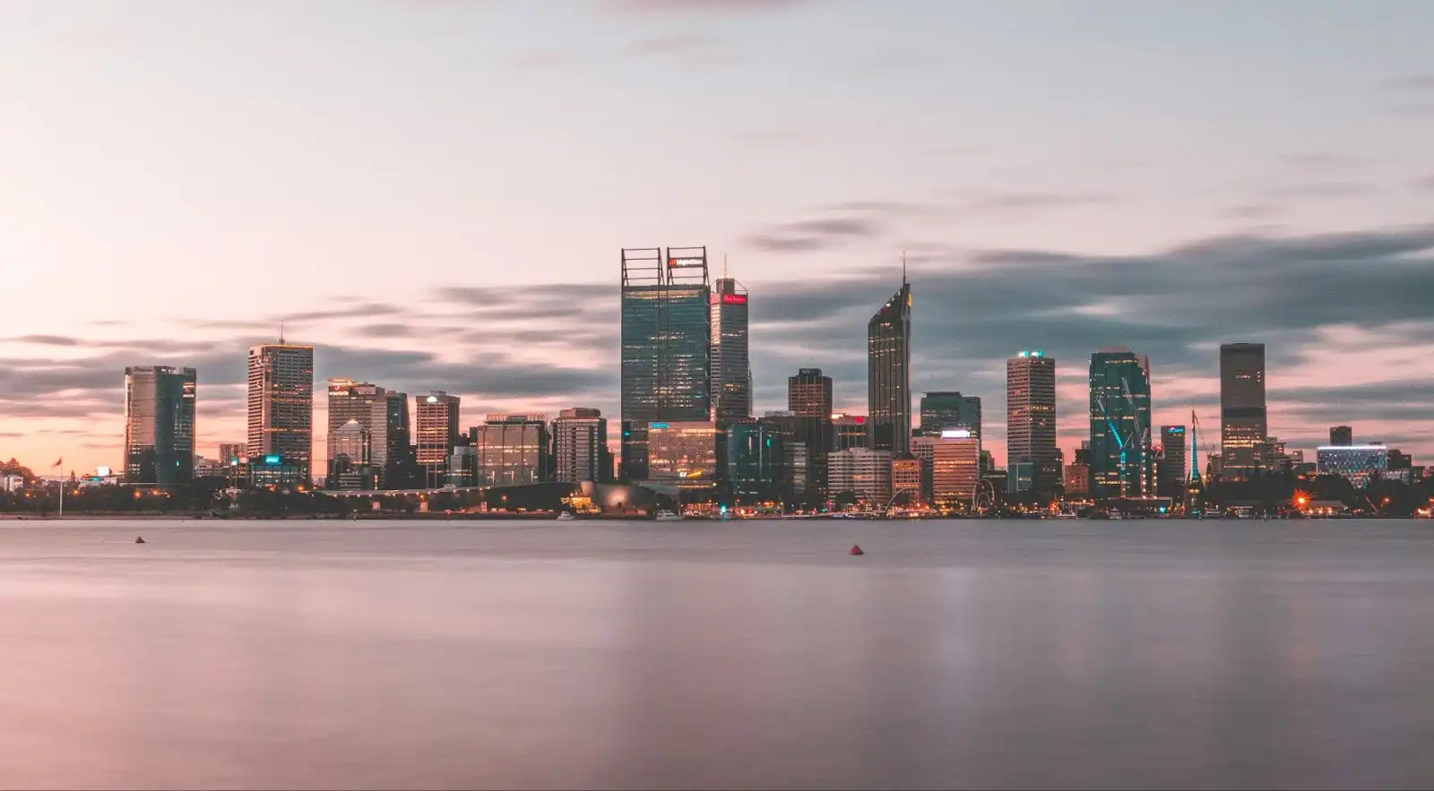 City skyline at dusk with skyscrapers reflecting in calm water, under a soft pink and blue sky. The scene conveys tranquility and urban beauty.