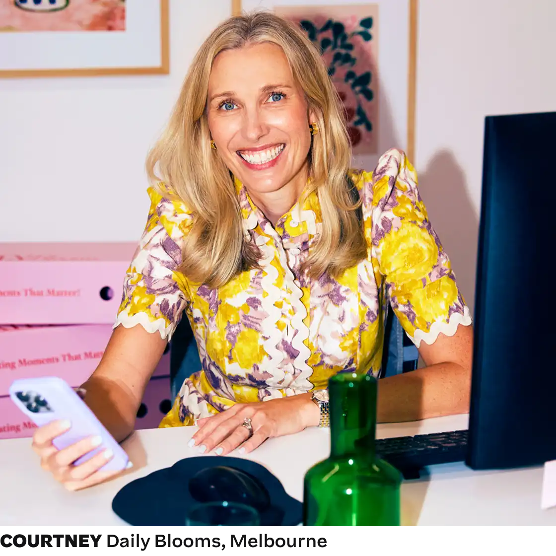 Smiling woman in a vibrant floral shirt holds a smartphone at a desk with a computer and green bottle, exuding a cheerful, professional vibe.