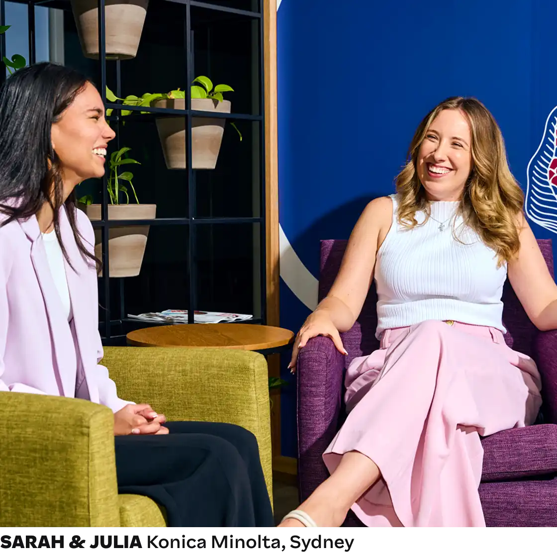 Two women laughing together in a modern office setting, one in a lavender blazer and the other in a white top and pink pants. The room has a vibrant blue wall and potted plants, creating a cheerful and relaxed atmosphere.