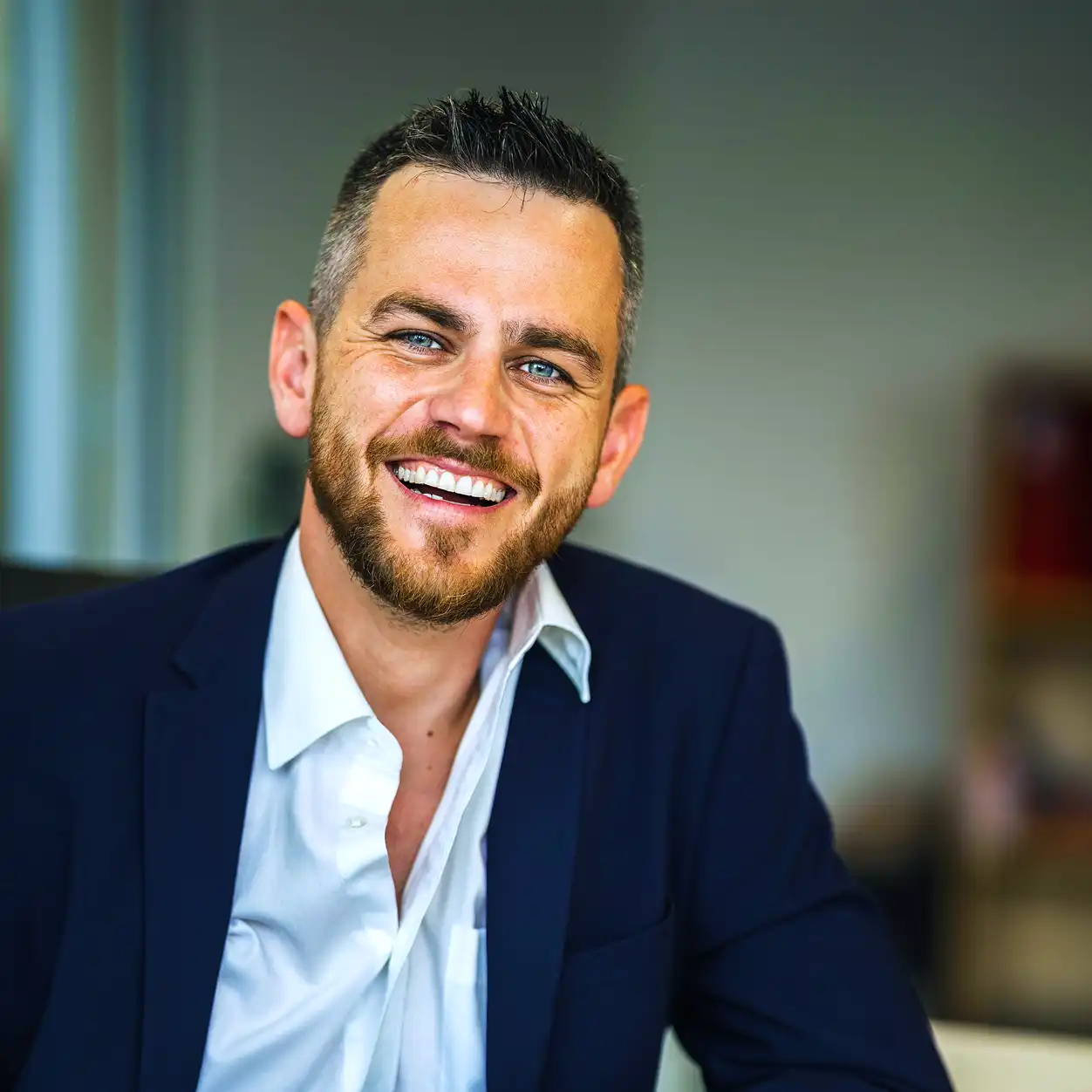 Smiling man with short hair and beard, wearing a dark suit and white shirt in an office setting. He conveys warmth and confidence.