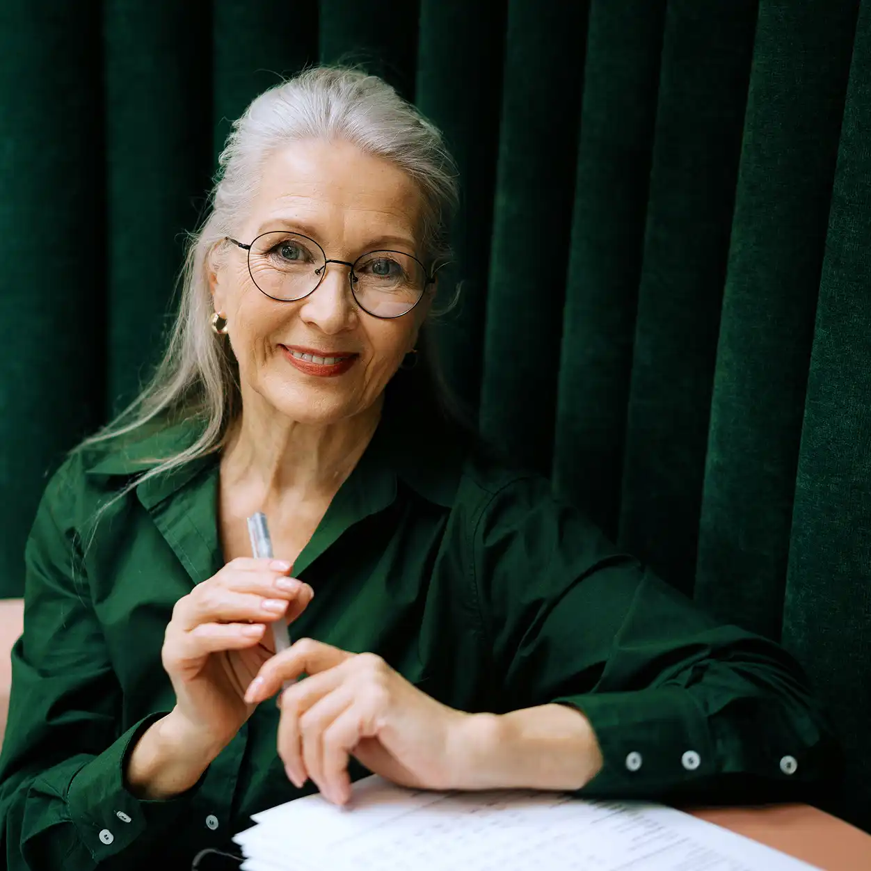 Elderly woman with glasses and long gray hair smiles warmly, holding a pen over papers, seated by green velvet curtains. She wears a dark green shirt.