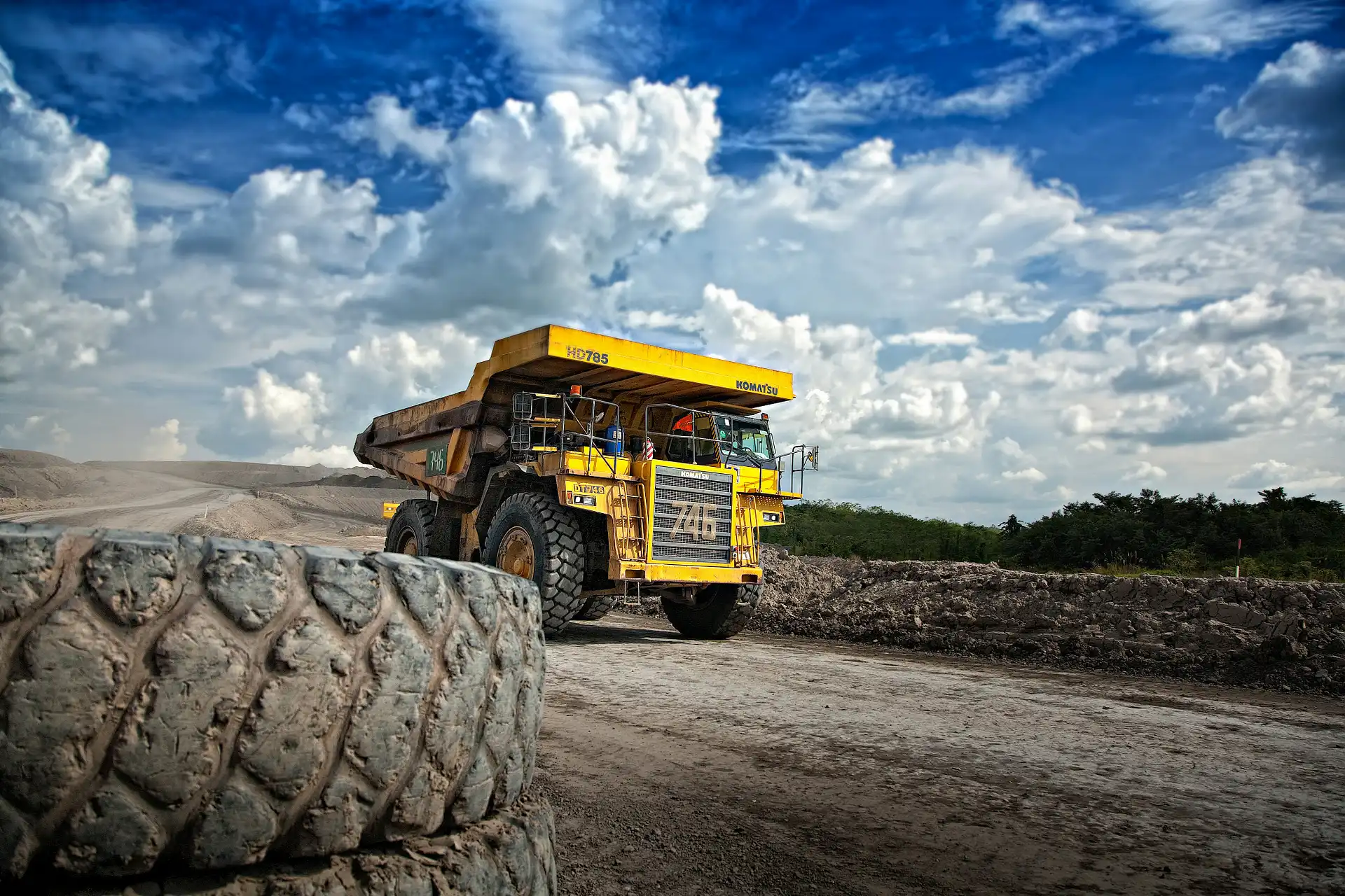 Yellow construction dump truck on a dirt road, with a large tire in the foreground. The sky is blue with fluffy white clouds. Rugged and dynamic scene.