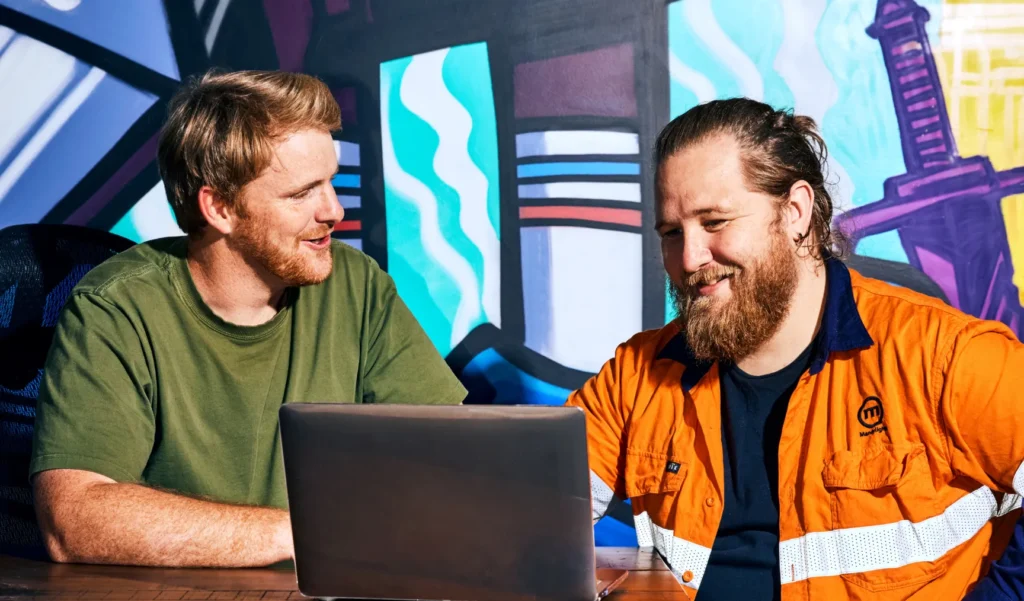 Two men smiling while working on a laptop in a creative workspace.