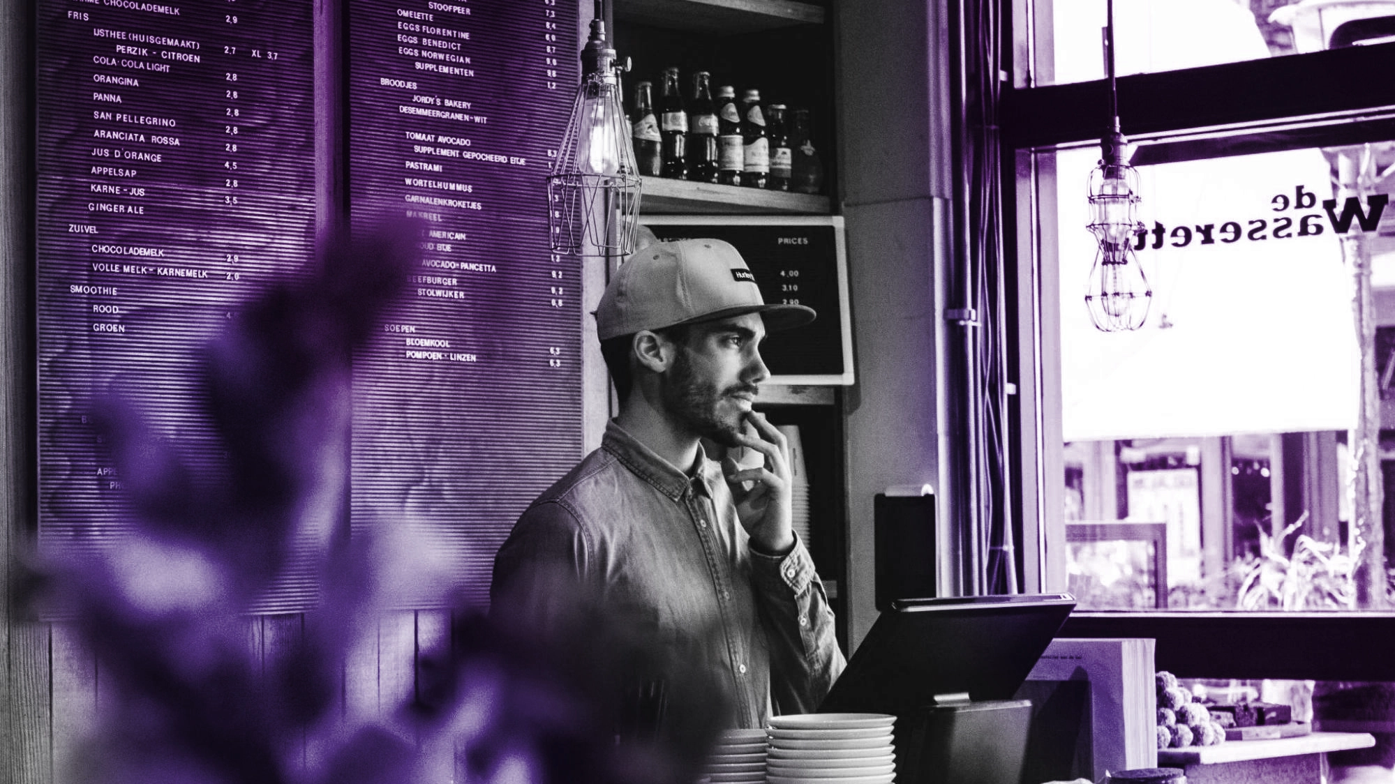 Barista standing behind café counter, looking thoughtful beside menu board.