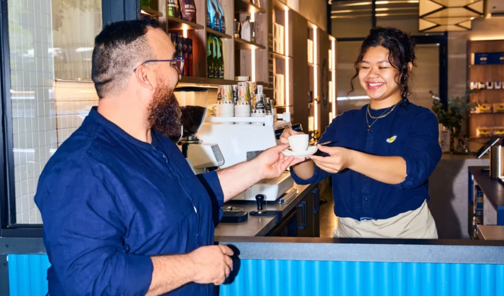 A smiling female barista standing behind a cafe counter handing a small cup of coffee to a happy male customer.