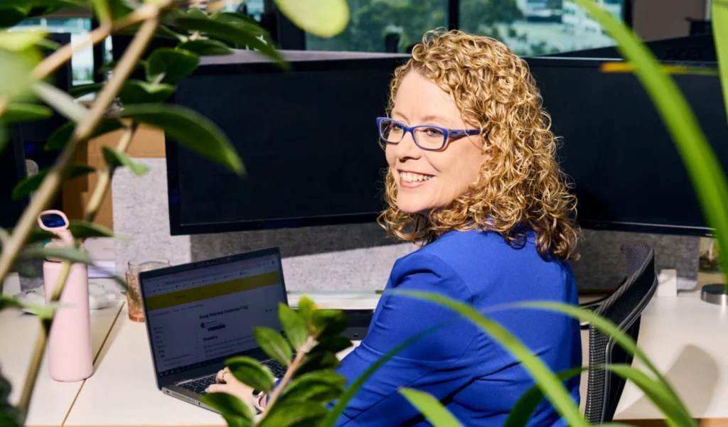 A smiling professional woman with curly blonde hair and glasses sitting at her office desk with a laptop and dual monitors, looking back over her shoulder.