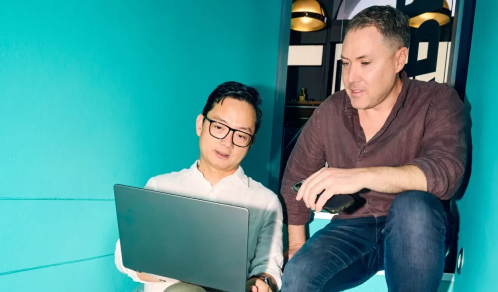 Two male colleagues sitting on bright teal stairs in a modern workspace, looking together at a laptop screen to discuss a project.