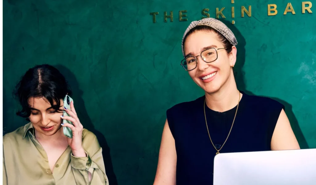 Two women are working in a store. One is on the phone, smiling, wearing a green shirt. The other, in glasses, smiles while sitting at a laptop. The mood is friendly.
