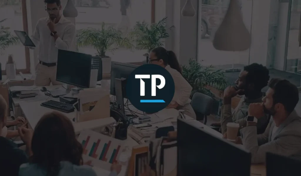 A diverse group of people in a modern office setting attentively listen to a presenter with a tablet. Green plants and large windows create a fresh, collaborative atmosphere.