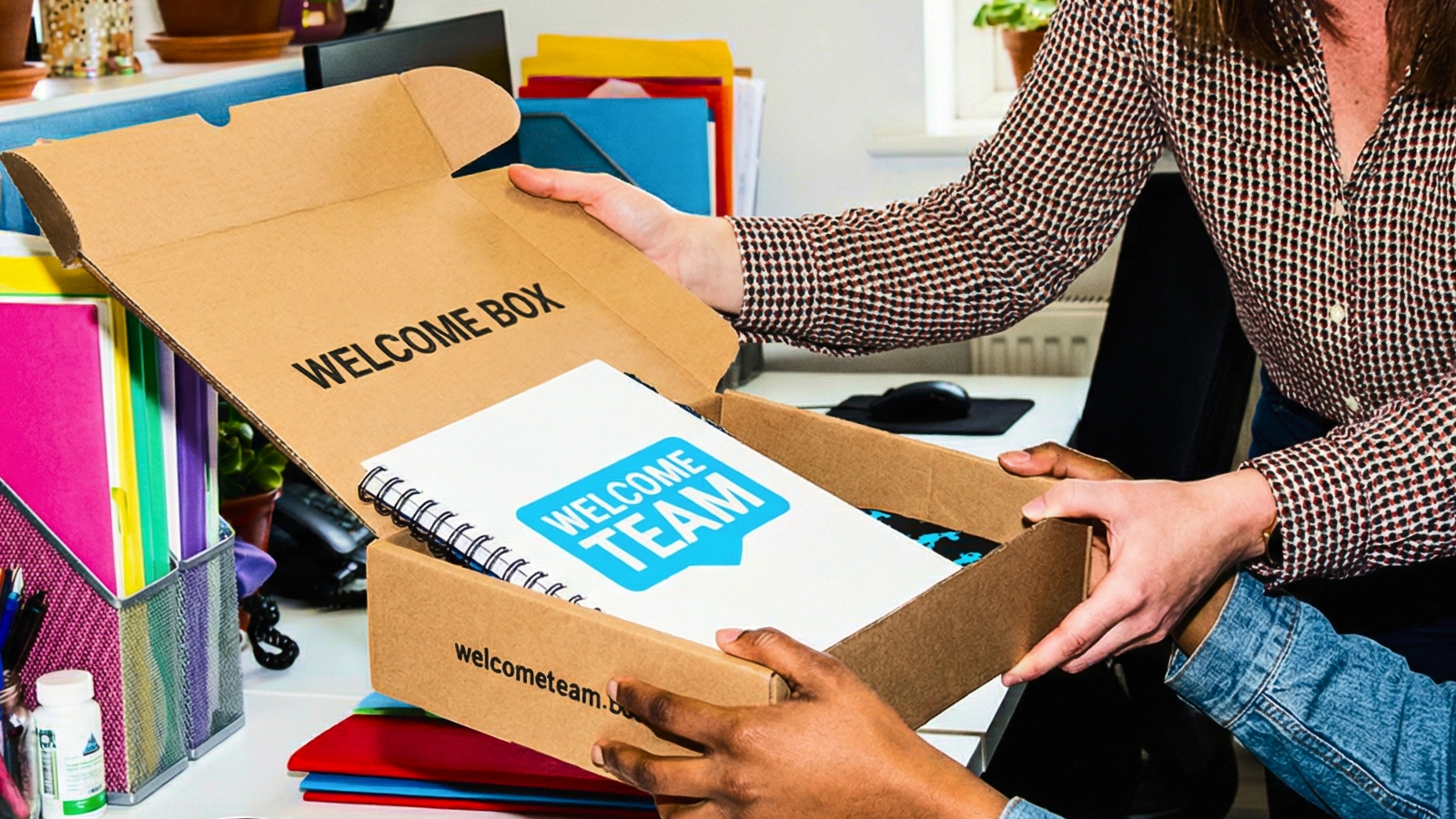 Hands opening a welcome box with a notebook labeled “Welcome Team” on an office desk.