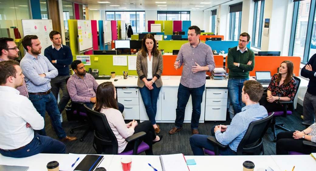 Team of office employees gathered in a modern open-plan workspace, listening as a man speaks during a group discussion.