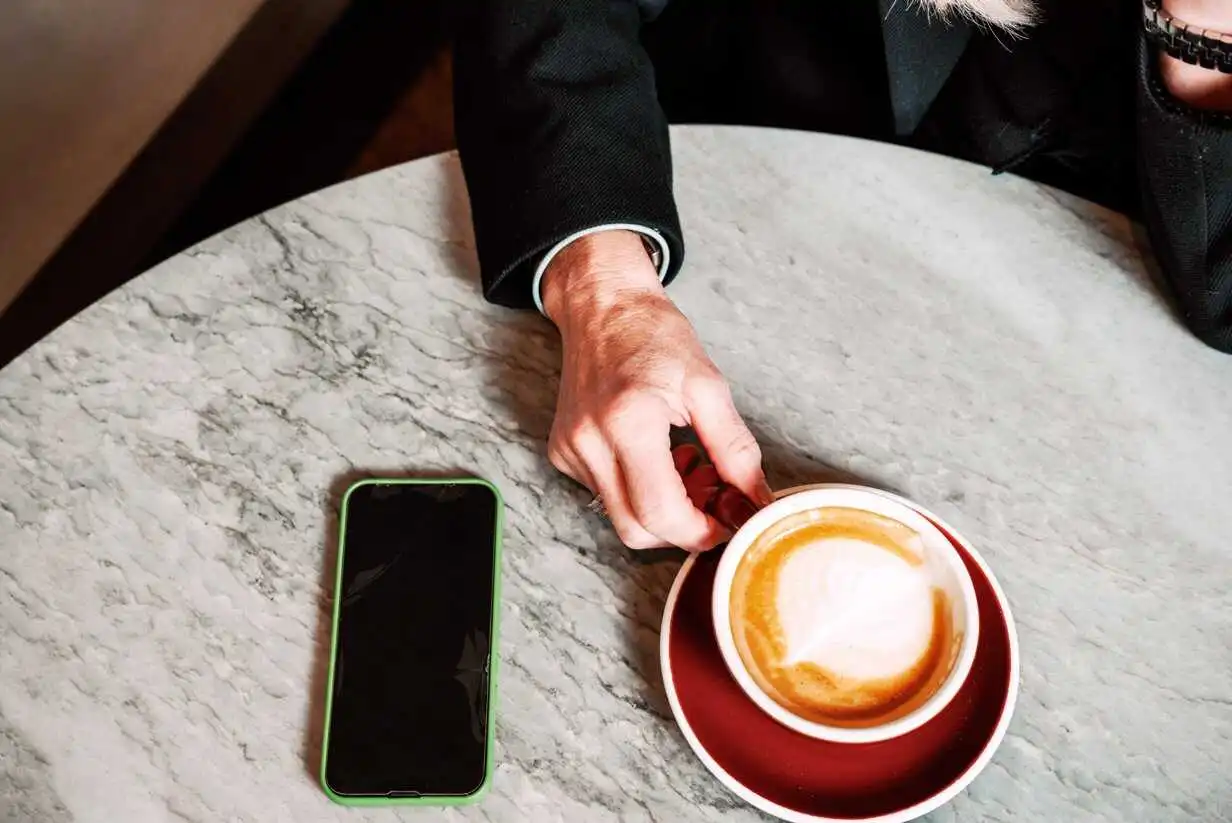 A hand holds a cappuccino in a red cup on a marble table. Nearby, a smartphone with a green case lies flat. The setting feels casual and relaxed.