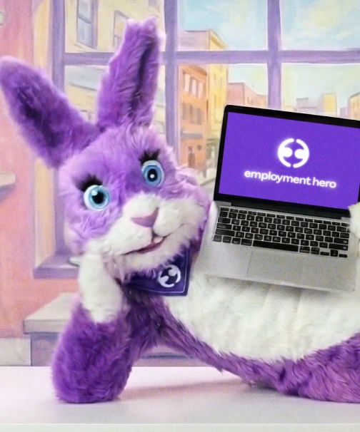 Purple plush rabbit with big blue eyes, casually resting its head on one hand. It holds a laptop displaying a purple screen with the "Employment Hero" logo. Background shows a colorful cityscape painting.