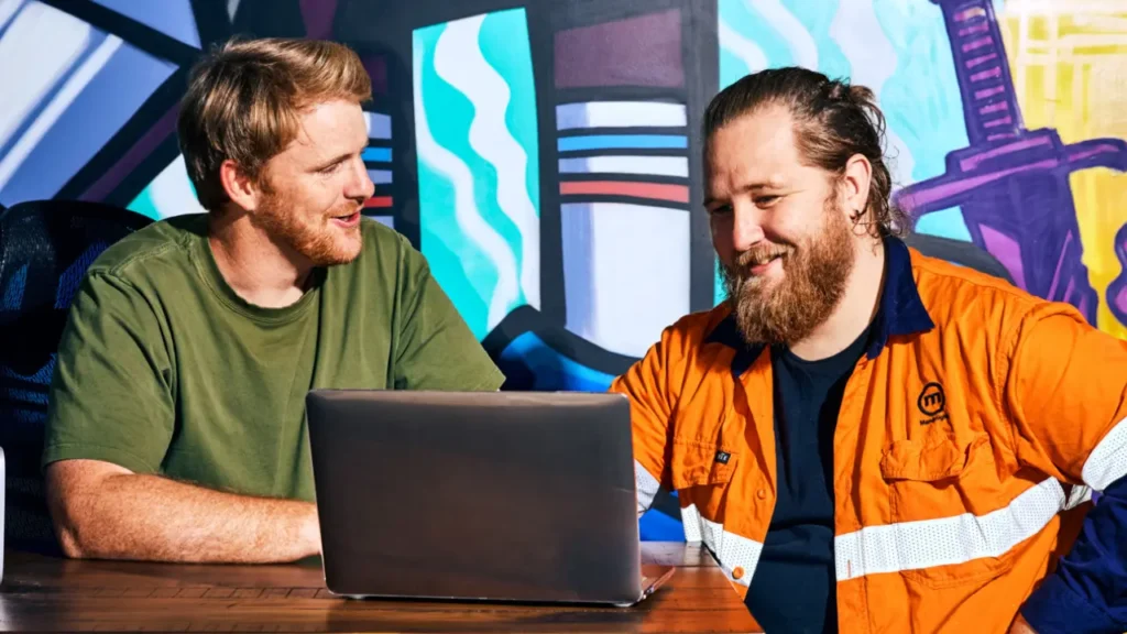 Two men, one in a green t-shirt and one in a high-visibility work shirt, looking at a laptop together.