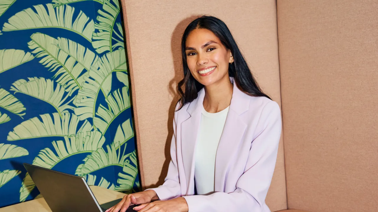 A smiling HR professional sitting with a laptop in a modern office booth, representing someone utilizing a talent acquisition audit checklist to improve hiring processes.