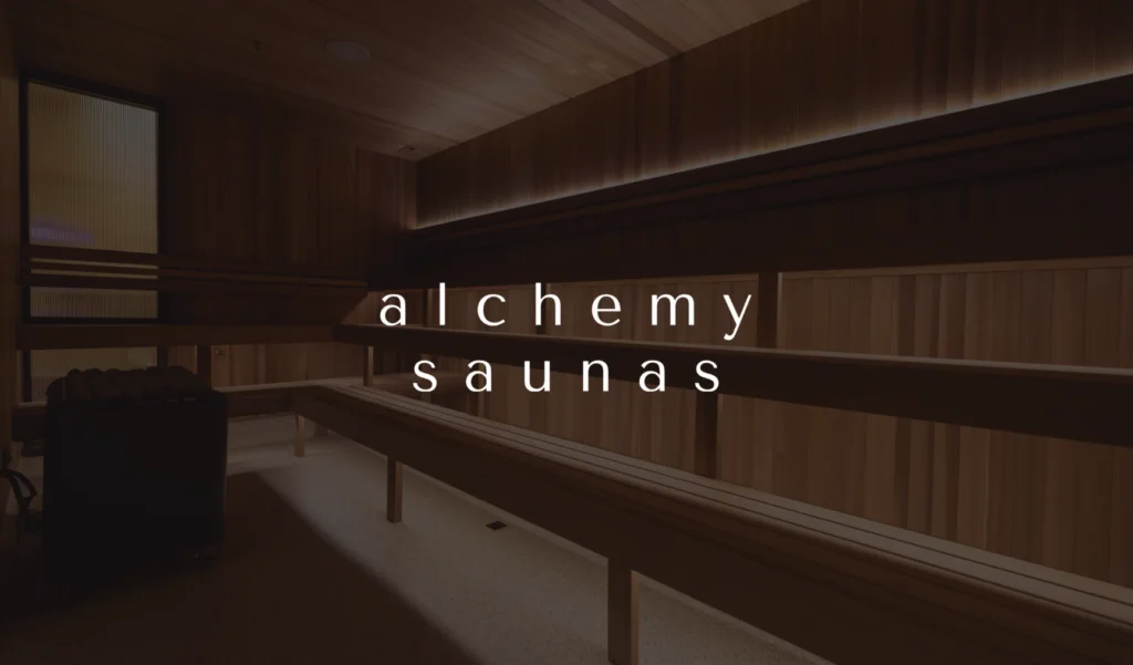 Dimly lit wooden sauna interior with benches, creating a warm and serene ambiance. The words "alchemy saunas" are centered in white text.