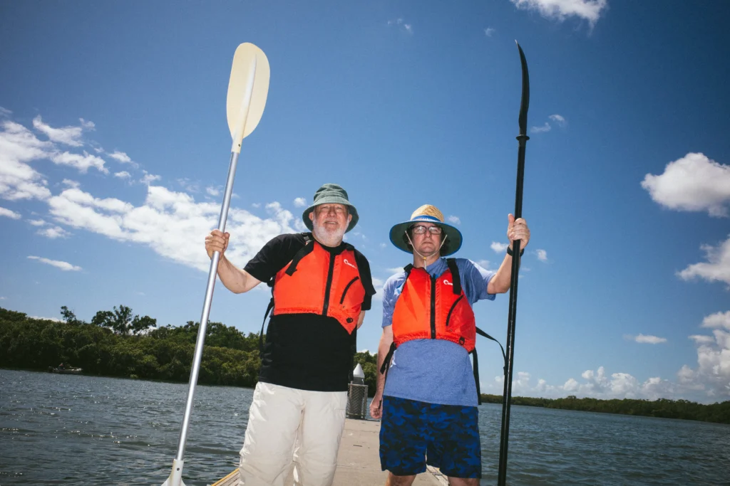Two men in orange life vests stand on a dock by water, each holding a paddle. They're wearing hats under a blue sky with fluffy clouds.