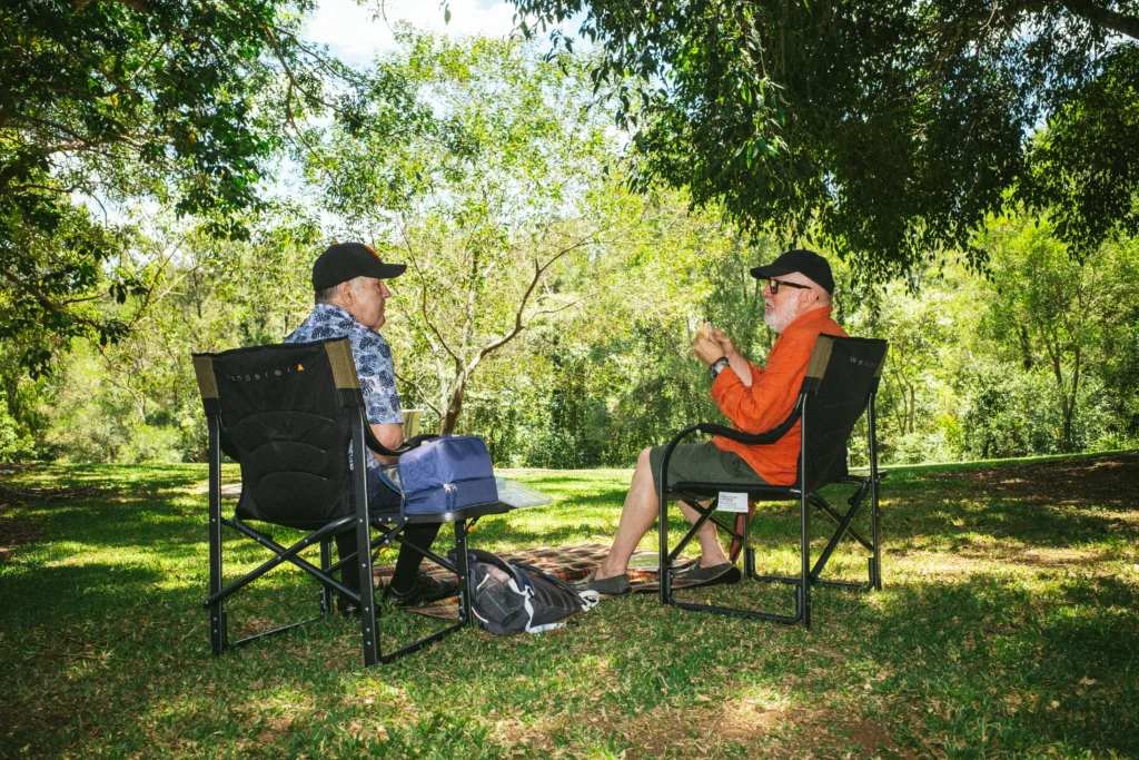 Two men sit on camping chairs in a lush, sunlit park, engaged in conversation. They are surrounded by greenery, exuding a relaxed, peaceful vibe.