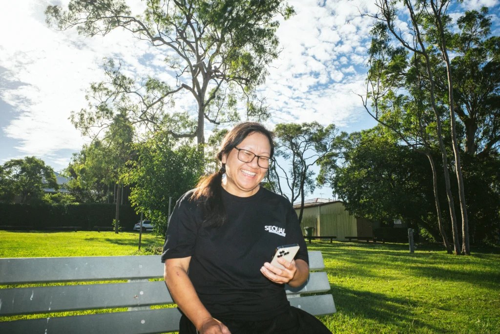 A joyful person sits on a park bench, smiling at a phone. They're wearing a black shirt. The park is sunny with scattered clouds and lush trees.
