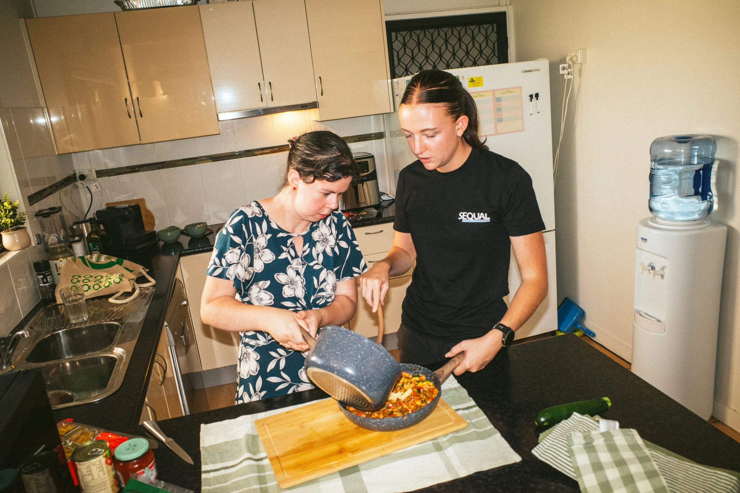 Two people in a kitchen pour food from a pot onto a plate. The kitchen has wooden cabinets, a refrigerator, and a water cooler, creating a cozy atmosphere.