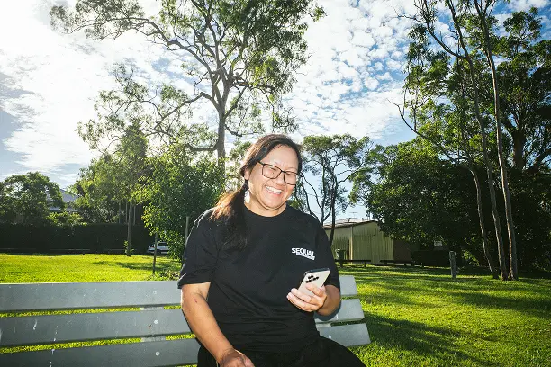A person sits on a park bench, smiling while looking at a smartphone. The sunny day and lush green trees convey a joyful, serene mood.