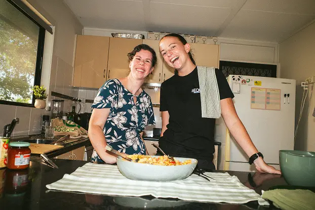 Two people smiling in a kitchen, standing behind a counter with a bowl of salad. A towel is draped over one shoulder, creating a casual, joyful atmosphere.