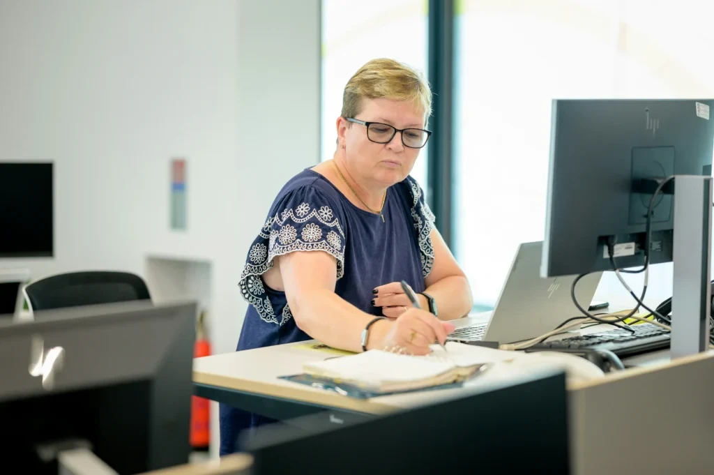 
A woman wearing glasses and a navy blue blouse writes in a notebook at a modern office workstation equipped with a laptop and a large computer monitor.
