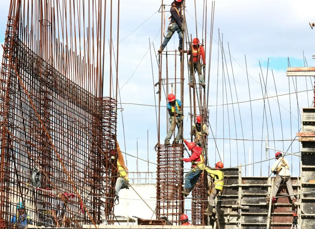 Several construction workers in hard hats and safety gear climbing and working on a tall steel rebar structure, representing the complex job sites where employers can utilize AI in construction for safety monitoring and workforce management.